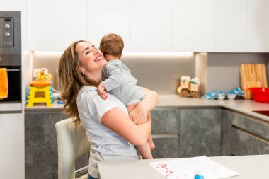 Anne bebeğini sevgiyle modern bir mutfakta kucaklıyor. Yürek ısıtan bir aile bağı yaratıyor.