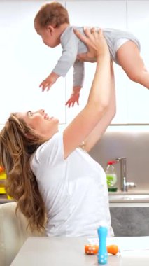 Sevecen anne neşeyle kaldırıyor, kucaklıyor ve bebeğiyle parlak mutfak ışığında etkileşime geçiyor. Gerçek bir annelik bağı ve neşeli bir aile anı sergiliyor.