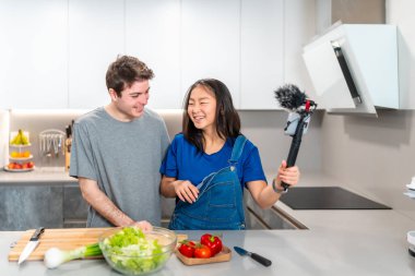 Mutlu çok ırklı çift salata hazırlıyor ve modern bir mutfakta yemek programı videosu çekiyor.