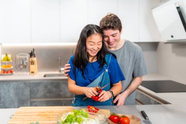 Çok ırklı bir çift evde yemek pişirmekten zevk alıyor. Parlak bir mutfakta ahşap bir tezgahta taze sebzeler hazırlarken gülümsüyordu.