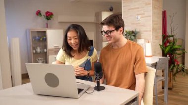 Laptop ve mikrofon kullanan çok ırklı bir çift modern dairelerinde podcast yapıyor.
