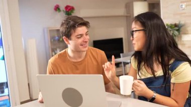 Genç, çok ırklı bir çift, birlikte rahatlatıcı bir sabah geçiriyorlar, dizüstü bilgisayarlarında geziniyorlar ve modern mutfaklarında kahve içiyorlar.