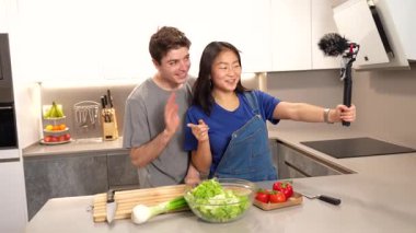 Çok ırklı çift video blogunda işbirliği yapıyor, ev mutfağında taze salata malzemeleriyle sağlıklı yemek hazırlıklarını kaydediyor.