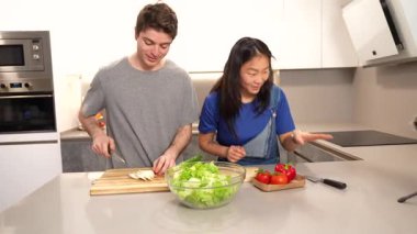 Çok ırklı çift, canlı, taze salatayı birlikte hazırlarken, sevgi dolu takım çalışması yaparken ve modern mutfak alanında ortak aşçılık becerileri paylaşırken iş birliği yapıyorlar.