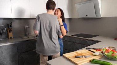 Genç çoklu etnik partnerler mutfak tezgahının yanında samimi bir şekilde dans ederken, sağlıklı yemek hazırlarken, modern ev ortamında neşe ve bağlantı yayarken, eğlenceli bir anı paylaşıyorlar.