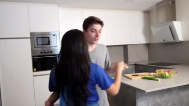 Genç çok ırklı çift, neşeli dans anlarını parlak, temiz, modern mutfakta paylaşıyor, spontane hareket ve yakın etkileşim yoluyla neşe ve bağlantılarını ifade ediyorlar.