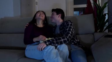 Genç, çok ırklı bir çift, oturma odalarında rahat bir kanepede otururken patlamış mısırlarını paylaşıyorlar.