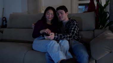 Çok ırklı bir çift kanepede dinleniyor, televizyon seyrediyor, uzaktan kumanda kullanıyor ve patlamış mısır yiyor.