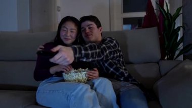 Mutlu, etnik çeşitlilikte bir çift evlerinde rahat bir akşamın tadını çıkarıyor, televizyon izliyor ve rahat koltuklarında patlamış mısır yiyor.