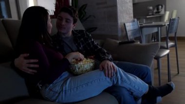 Çok ırklı bir çift, rahat koltuklarında patlamış mısırlarını ve sevgilerini paylaşarak evde rahatlatıcı bir akşamın tadını çıkarıyor.