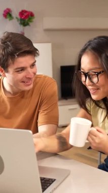 Genç çok ırklı çift, kahvenin keyfini çıkarırken ve modern dairelerinde dizüstü bilgisayar kullanırken neşeli bir anı paylaşıyor.