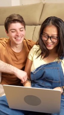 Çok ırklı bir çift, birlikte internet gezerken, rahat bir koltukta otururken ve parlak oturma odasında dizüstü bilgisayarla iletişim kurarken mutlu anları paylaşıyor.