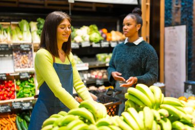 Süpermarkette müşteriyle konuşurken tezgahta taze muz düzenleyen manav.