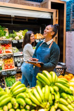 Gülümseyen market tezgahtarı elinde dijital tabletle müşteriye ürünleri gösteriyor, organik gıda süpermarketi