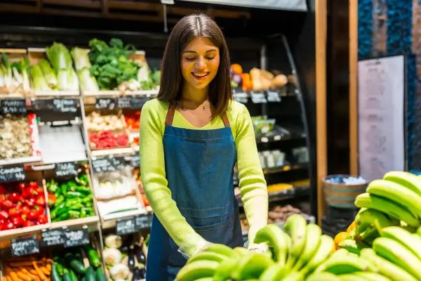 Tezgahta taze muz düzenleyen, meyve ve sebzeli organik süpermarkette çalışan gülümseyen bir satıcı kadın.
