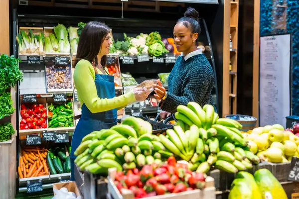 Dost canlısı bir satıcı markette taze meyve ve sebzelerle dolu raflarla bir müşteriye paketlenmiş bir ürün veriyor.