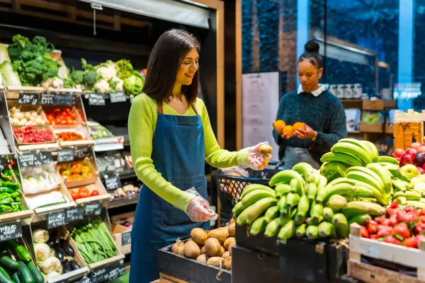 Ekranda taze kivi meyvesi düzenleyen eldiven takan süpermarket çalışanı, müşteri arka planda portakal seçerken,