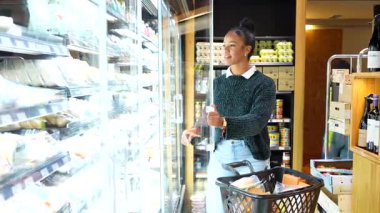 Kadın alışverişçi soğutulmuş rafları karıştırıyor, modern marketin içinde alışveriş sepeti tutarken taze organik ürünleri dikkatlice seçiyor.