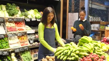 Eldiven takan süpermarket çalışanı muz ve çileklerin yanında patates düzenlerken, müşteri portakal seçiyor.