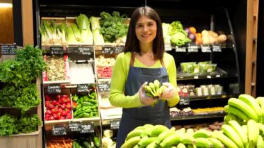 Marketteki tezgahtar eldiven takmış süpermarketin içinde yeşil muzlar sunuyor, kameraya gülümsüyor.
