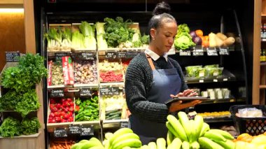 Dijital tablet kullanan bir süpermarket çalışanı, organik bir markette taze ürünler seçen bir müşteriye yardım ediyor.