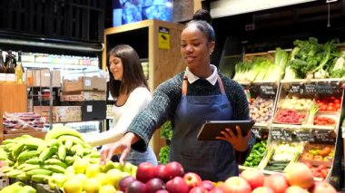 Profesyonel satıcı, markette renkli ürünlerin yanında dururken müşteriye taze, olgun organik meyveler seçmesinde yardımcı olmak için dijital tablet kullanıyor.