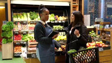 Süpermarket çalışanı bakkal alışverişinde müşteriye yardımcı oluyor, organik meyve ve sebzeler hakkında bilgi sağlıyor