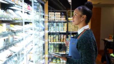 Market çalışanı envanteri yönetiyor, son kullanma tarihlerini kontrol ediyor ve soğutmalı vitrinde ürünleri düzenliyor.