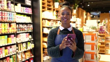 Akıllı telefon kullanan, gülümseyen ve kameraya bakan bir süpermarket çalışanı, organik ve glütensiz ürünler üzerine uzmanlaşmış bir markette çalışıyor.