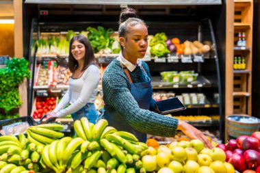 Market çalışanı elinde tablet tutarken taze meyve düzenlerken bir müşteri ve arka planda organik ürünlerle dolu raflar tutuyor.