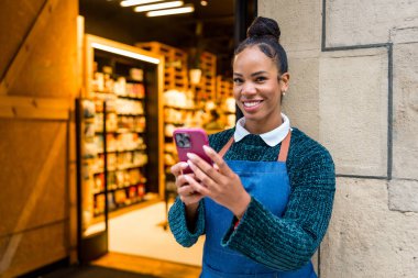 Mutlu Afro-Amerikan satış elemanı önlük giyiyor organik süpermarket girişinde akıllı telefon kullanıyor.