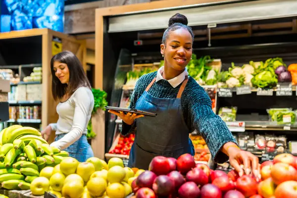 Gülümseyen satıcı kadın taze elmaları düzenlerken ve dijital tablet kullanırken müşteri süpermarkette muz seçiyor.