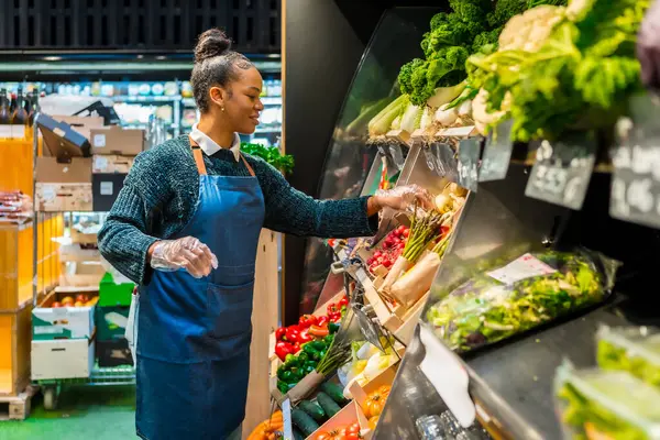 Süpermarkette organik sebzeleri düzenleyen eldivenler giyen bir market çalışanı.
