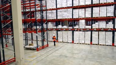 Turuncu yelek ve beyaz kask giyen lojistik mühendisi raflarla dolu büyük bir depoya giriyor.