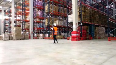 Profesyonel lojistik operatörü, geniş bir deponun içini geziyor. Yükselen raflar arasında ticaret ve endüstriyel depolama malları taşıyor.