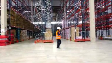 Güvenlik yeleği ve kask giyen kadın müfettiş büyük dağıtım merkezindeki raflarda mal ve malları denetliyor.