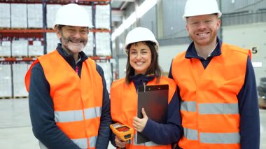 Depo lojistik uzmanları parlak güvenlik teçhizatı ve kasklar takıyor, organize endüstriyel depolama tesisinde çalışırken güvenle gülümsüyorlar.