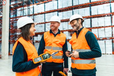 Güvenlik yelekleri ve şapkalar giyen depo çalışanları büyük bir dağıtım merkezinde barkod tarayıcıları kullanarak gülümsüyorlar.