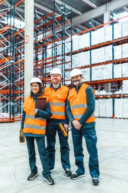 Depoda güvenlik yelekleri ve kask takan işçiler büyük bir depoda envanter yönetim aletlerini tutarken gülümsüyorlar.