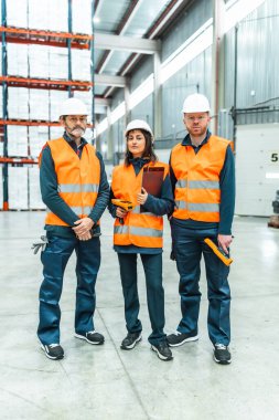 Güvenlik yelekleri ve kasklar takan depo işçileri büyük bir dağıtım merkezinde envanter ve lojistik işlerini yürütüyorlar.