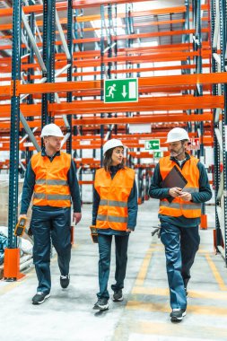 Güvenlik yelekleri ve başlık takan depo çalışanları büyük bir dağıtım merkezinden geçerek lojistik ve envanter yönetimini tartışıyorlar.