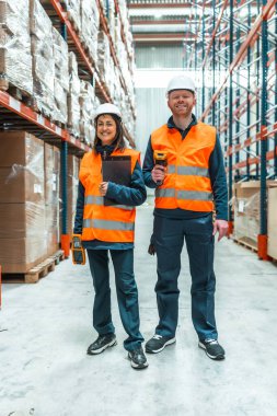 İki depo işçisi envanteri yönetiyor, ellerinde barkod tarayıcı ve dosya var, büyük bir dağıtım merkezinde gülümsüyorlar.