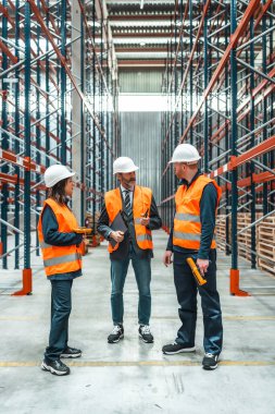 Depo çalışanları ve yöneticileri büyük bir dağıtım merkezinde rafların arasında dururken lojistik operasyonları tartışıyorlar.