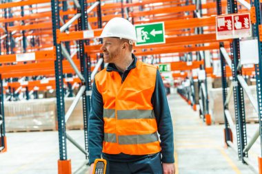 Güvenlik yeleği ve kask giyen bir depo işçisi lojistik merkezine doğru yürüyor ve gülümsüyor.
