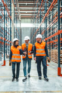 Güvenlik yelekleri giyen depo çalışanları ve yöneticileri büyük bir dağıtım merkezinde yürüyorlar ve lojistiği tartışıyorlar.