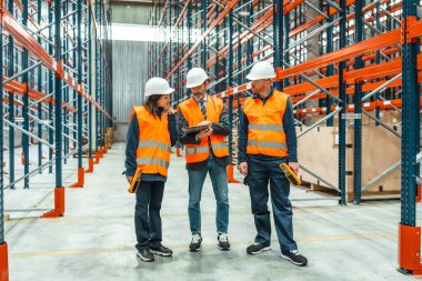 Depo çalışanları ve yönetici, büyük bir dağıtım merkezinde raflarla çevrili dijital tablet kullanarak lojistiği tartışıyorlar.