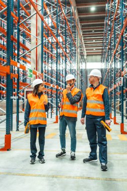 Güvenlik yelekleri ve miğferler giyen depo çalışanları büyük bir dağıtım merkezinde telsiz ve barkod tarayıcısı kullanarak lojistiği tartışıyorlar.