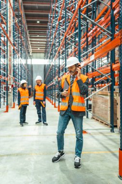 Telefon ve pano kullanarak lojistik operasyonlarını koordine eden yönetici, depo ortamında işbirliği yapan yüksek görünürlük yelekleri giyen işçiler