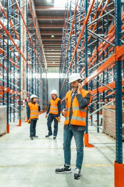 Depo müdürü güvenlik yeleği ve kask takıyor akıllı telefon kullanıyor, büyük dağıtım merkezindeki ekibiyle lojistik operasyonları koordine ediyor.