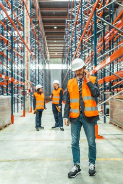 Depo müdürü güvenlik yeleği ve kask takıyor cep telefonu kullanıyor, büyük dağıtım merkezindeki ekibiyle lojistik operasyonları koordine ediyor.